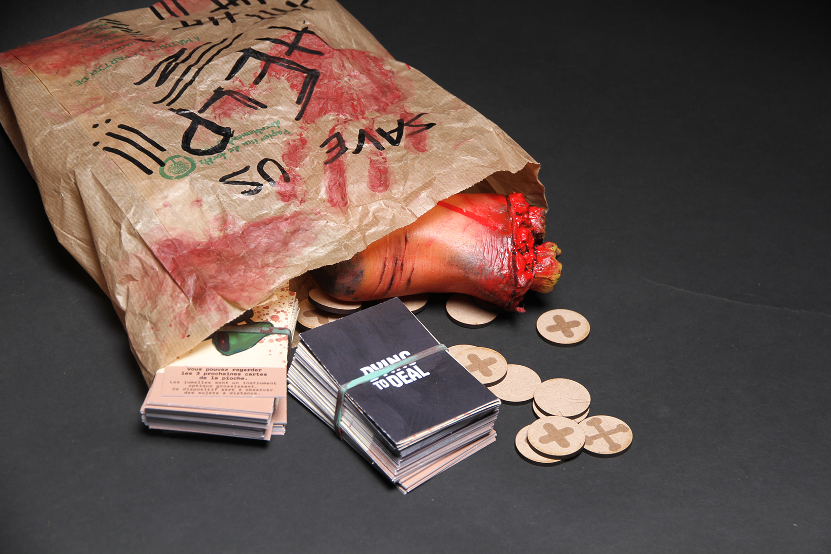 photographie d'un sac en papier qui contient un pieds en plastique, deux paquets de cartes, et des jetons. On peut lire save us et help sur le sac.