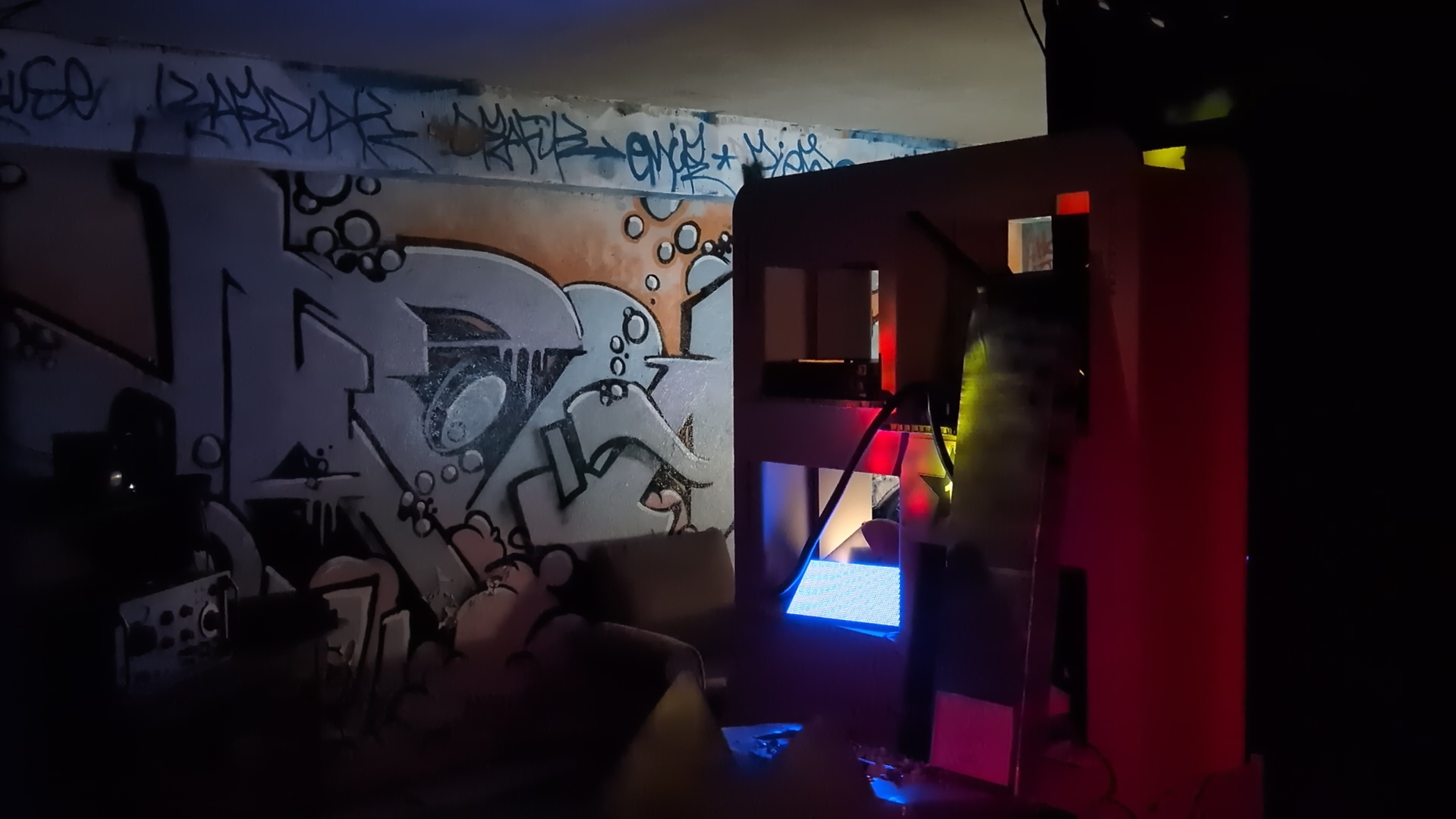 Photographie d'un lieu sombre éclairé par des lumières de différentes couleurs, dans lequel on voit des structures de cartons et des vieux outils électroniques