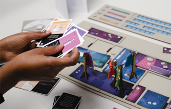 photographie d'une personne qui tient un éventail de cartes illustrées colorées, à côté d'un plateau de jeu modulaire